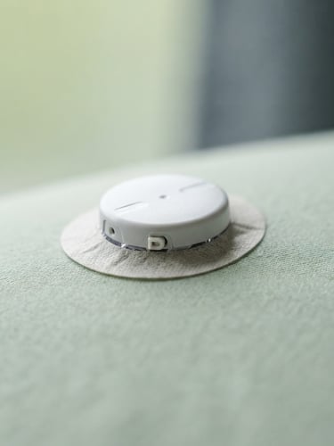 Macro shot of a modern continuous glucose monitor sensor on a clean, light sage green surface, soft natural daylight, shallow depth of field