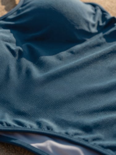 Close-up macro shot of premium textured swimwear fabric, rich ocean blue weave catching warm golden-hour sunlight, showing fine details of the high-performance lycra, shallow depth of field.