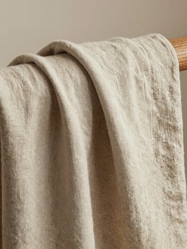 A portrait crop of a neutral sandstone-colored linen fabric draped elegantly over a wooden rail, highlighting the soft tactile weave under diffused natural light.