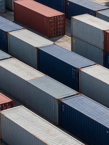 Overhead geometric pattern of multi-colored shipping containers stacked precisely in an automated terminal, sharp morning light, high contrast.