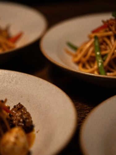 Close-up macro shot of textured ceramic plates with a vibrant Asian fusion dish, dramatic low-key side lighting, warm amber tones, extreme shallow depth of field, 35mm