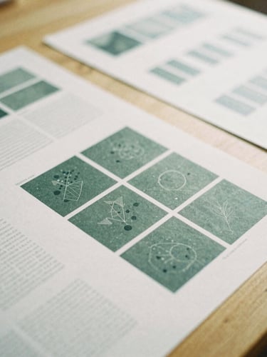 Close-up of physical design proofs and editorial layouts laid out on a clean wooden table, soft overcast British daylight, 35mm film grain