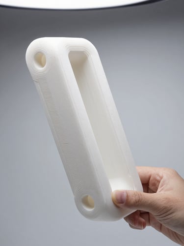 Side-profile close-up of a complex white 3D-printed mechanical bracket held at arm's length against a neutral light grey studio background, showing crisp layer lines and support removal marks, studio-bright even overhead light, depth of field on the surface detail at 2cm