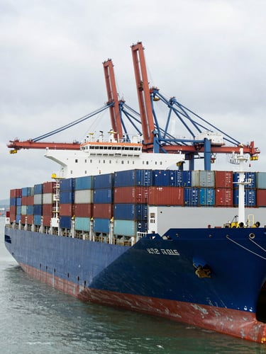 Wide environmental shot of a massive container ship docked at Port Klang under overcast maritime skies, stacked with blue and red containers, heavy harbor cranes in the background.
