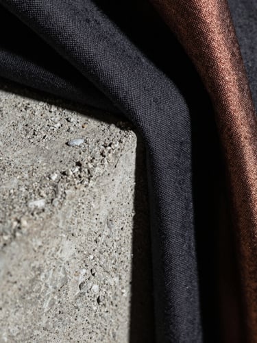 Macro photograph of rough-hewn raw concrete meeting heavy, dark, hand-stitched textile fabrics, high-contrast side lighting with deep shadows and metallic copper highlights.