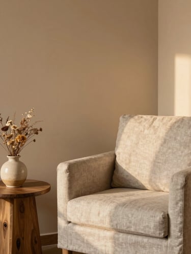 A cozy, empty corner of a modern psychotherapy room. A soft linen armchair, a small ceramic vase with dried wildflowers on a wooden side table, warm diffused golden-hour light casting soft shadows on a warm sand-colored wall.