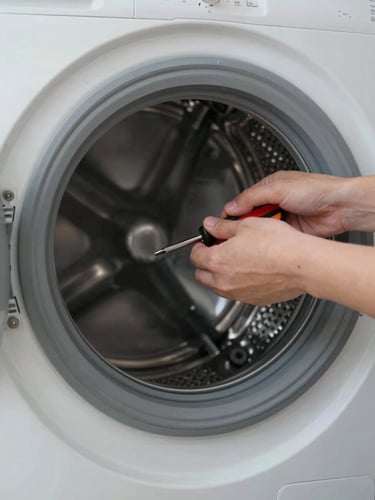 Close-up of a technician's hands removing the drum panel of a front-load washing machine, bright natural window light from the left, screwdriver visible mid-action, laundry room context in soft focus behind