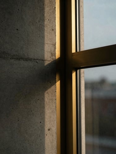 Close-up architectural crop of raw board-formed concrete meeting a brushed brass window frame, moody overcast daylight, shallow depth of field, rich desaturated tones.