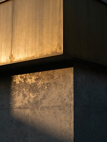 A close-up architectural photograph of raw concrete meeting brushed brass under low golden-hour light, casting long dramatic shadows.
