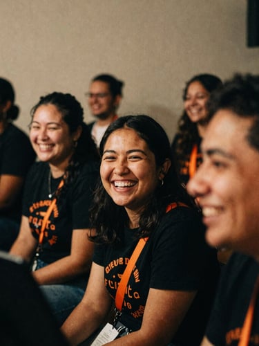 Close-up, slightly off-kilter, gritty film photography of diverse, smiling volunteers sharing a laugh while setting up for a concert in a dimly lit community space, warm beige and deep black tones, vibrant orange accents, 35mm grain.