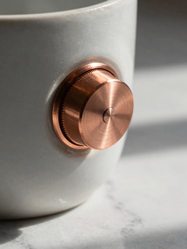 A close-up macro photograph of a brushed-copper weighted dial on a matte ceramic vessel, soft morning light casting long shadows across a clean marble countertop.
