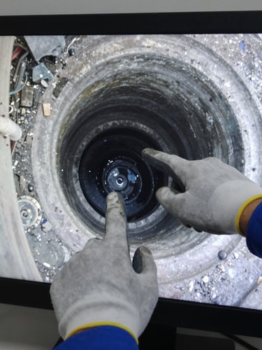 Close-up of a CCTV monitor screen displaying live pipe footage in natural daylight, technician's gloved hand pointing at a visible blockage on screen, equipment cables in foreground