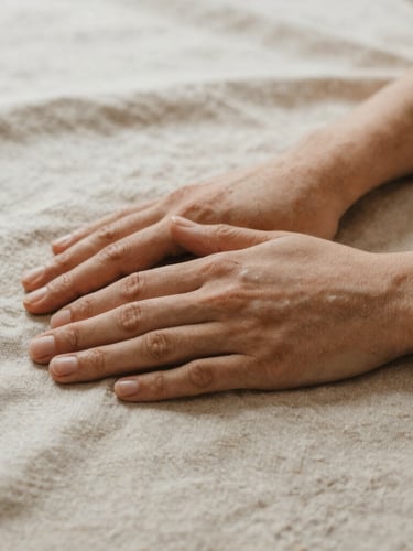 Close-up of hands gently resting on a textured organic linen fabric, soft natural overcast daylight, warm sand tones, shallow depth of field, minimalist somatic aesthetic.