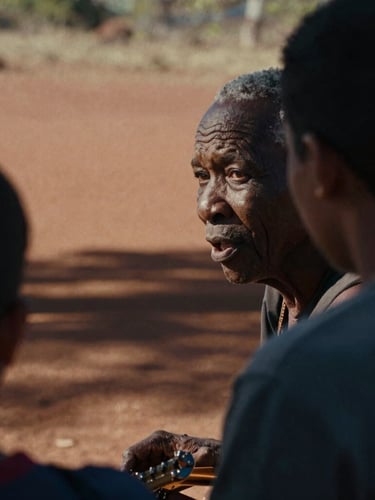 An intimate close-up of an Aboriginal Elder speaking with a young musician in a sunlit outdoor setting, warm red dirt in the background, high-contrast natural light, cinematic 35mm film grain.