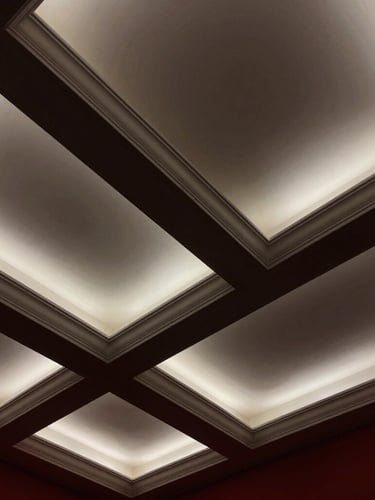 Low-angle architectural photograph looking up at a pristine, geometric white coffered ceiling with integrated warm lighting, dramatic raking light highlighting crisp millwork joints, high-contrast charcoal shadows, 35mm lens.