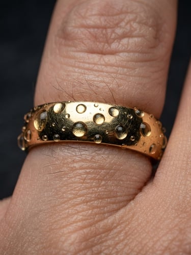 Extreme macro shot of a solid 14k gold ring on skin, water droplets clinging to the metal, high-contrast studio lighting, deep charcoal background