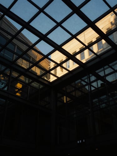 Dramatic architectural perspective looking up at a modern glass ceiling with golden hour reflections, low-key lighting, deep black shadows, 35mm