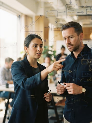 Candid portrait of two business professionals analyzing structured diagrams on a glass board, warm natural window light, deep navy and soft gold tones, shallow depth of field, 35mm