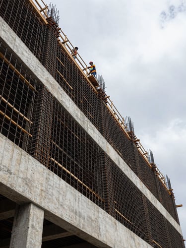 Wide architectural documentary shot from ground level looking up a reinforced concrete shear wall under construction in São Paulo, steel rebar grid exposed mid-pour, overcast diffused morning light eliminating shadows, workers in safety helmets visible at upper scaffold level, structural geometry filling the left two-thirds of the frame