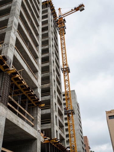 Wide environmental shot from ground level looking up a residential building under active construction in São Paulo, concrete formwork and rebar columns rising into an overcast morning sky, construction workers and crane visible at mid-frame, strong vertical geometry dominating the left edge of the frame