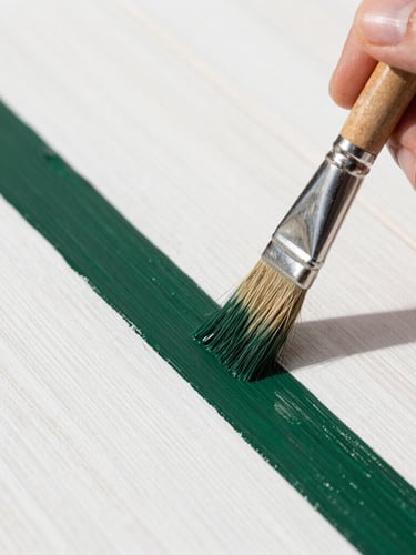 Close-up macro shot of a hand holding a high-quality paint brush applying a perfectly straight line of dark green paint onto a crisp white wooden surface, bright natural daylight, sharp focus.