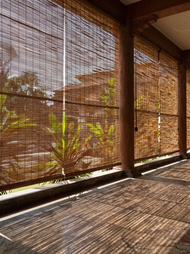 A grand hotel verandah in Lucknow at midday, featuring massive hand-woven bamboo blinds filtering intense sunlight into beautiful linear shadows on a polished marble floor, lush green palms in the background.