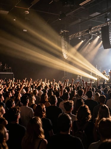 Wide environmental shot of a packed indoor concert crowd under stage lighting, golden beams cutting through dark air, audience energy visible in motion, shot from slightly elevated stage-right angle, cinematic high-contrast