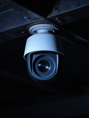 Cinematic portrait shot of a high-end security camera lens mounted on a dark industrial steel beam, dramatic high-contrast night lighting, sharp focus on the glass lens, cool blue ambient glow, 35mm