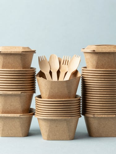 Straight-on product shot of neatly stacked, uniform brown kraft paper takeout containers and compostable wooden cutlery, bright high-key studio lighting, neutral slate-blue background.