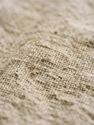Macro detail of raw flax linen texture, soft natural window light highlighting the organic weave and subtle wrinkles, warm oatmeal and raw plaster tones, shallow depth of field, editorial 35mm film photography