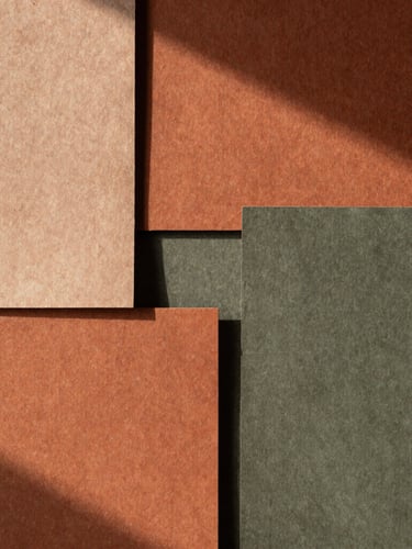 A tall portrait shot of textured handmade paper swatches in warm terracotta and muted olive tones, soft directional afternoon light casting deep shadows.