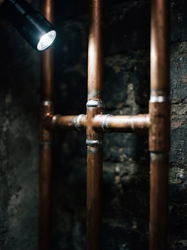 Tight portrait macro shot of clean copper piping joints with fresh solder, illuminated by a sharp utility flashlight beam against a dark charcoal background, high-contrast orange and charcoal tones, 35mm