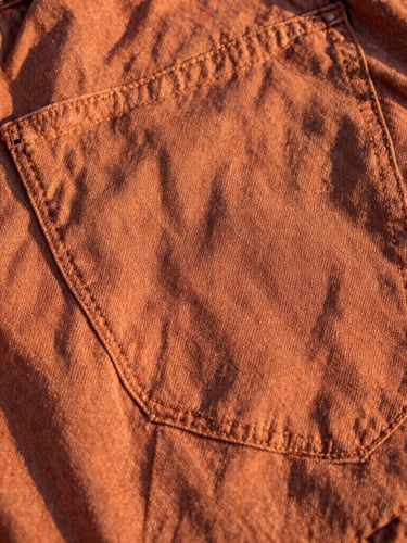 Close-up of worn terracotta-toned denim fabric showing beautiful textured fade lines and sturdy double-stitching in warm golden-hour sunlight.