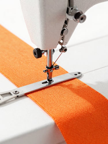 Close-up of industrial sewing machine stitching heavy-duty orange webbing, bright high-key daylight, metal buckles nearby, sharp focus