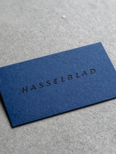 Macro close-up of premium heavy-weight cotton business cards with deep debossed black typography on a cool grey concrete surface, sharp studio lighting, minimal composition