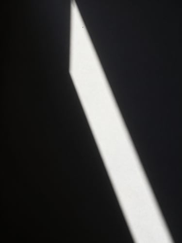 Narrow vertical frame, a single shaft of direct sunlight cutting diagonally across an otherwise pitch-dark interior wall, the boundary between light and shadow razor-sharp, monochrome tones, high contrast, no subject other than light itself