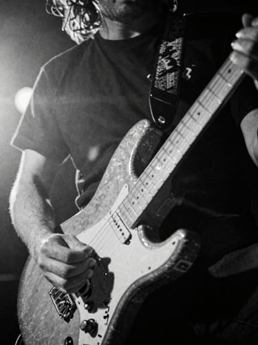 Extreme close-up, low-angle, high-contrast black and white shot of a guitarist's hand shredding on a battered electric guitar, stage lights creating lens flares, motion blur, 35mm film grain, bleeding right.