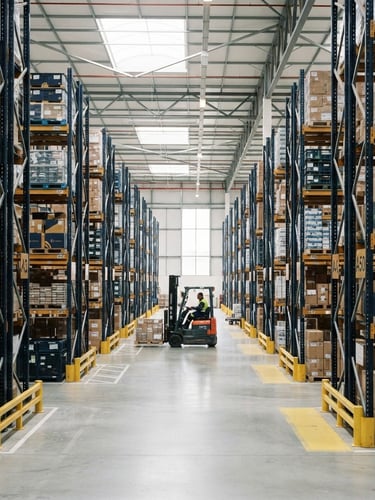 Wide shot of a modern, well-lit logistics warehouse interior with high shelves, organized inventory, and a forklift in the distance, professional corporate photography, bright natural daylight, clean navy and gold accents, sharp focus, architectural framing, 35mm