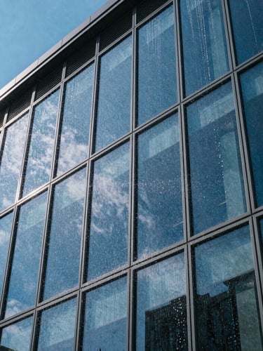 Architectural angle of a modern glass facade with sharp reflections of a clear blue sky, water sheeting cleanly off the pristine glazing surface during a low-impact wash.