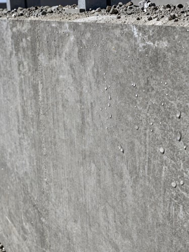 Extreme close-up of water-beading on a freshly sealed concrete foundation wall under high-contrast midday sun