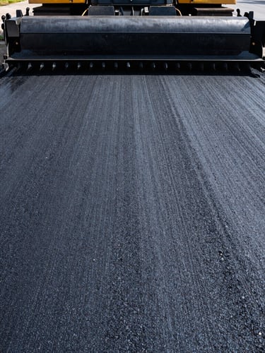 Low-angle wide shot of a heavy-duty asphalt paving machine on a fresh black surface under bright midday sun, deep shadows, gritty industrial texture.