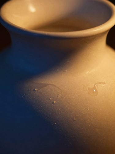 Low-key warm golden candlelight casting deep blue shadows over an elegant ceramic vessel, macro texture of melted wax, shallow depth of field.