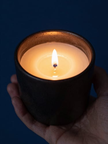 Macro close-up of a hand gently holding a dark, textured ceramic candle vessel, warm golden candlelight reflecting off the smooth wax, deep blue shadow background.