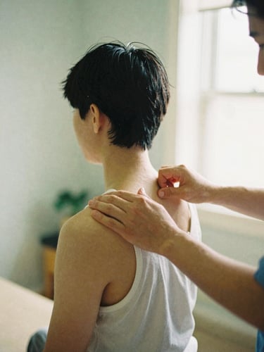 Editorial 35mm film photo of a physical therapist's hands gently adjusting a patient's shoulder in a bright, naturally lit Elm Grove clinic.