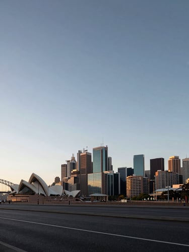 Wide panoramic view of Sydney CBD skyline at early morning, glass towers catching cool pre-dawn light, low horizon with the harbour bridge edge visible far left, vast grey-blue sky dominating the upper two-thirds of the frame, empty foreground street