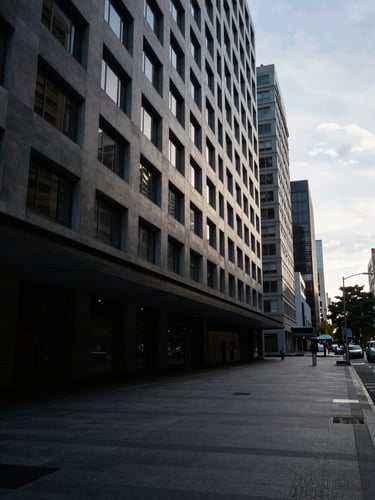 Wide environmental shot of a mid-rise commercial streetscape in a major Australian CBD at early morning, cool overcast sky, empty footpath, building facades in deep shadow with pale grey light catching upper floors, no people visible