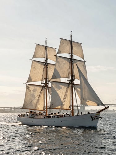 Wide environmental shot of a classic 80-foot sailing yacht underway on Narragansett Bay, crisp morning light reflecting off the silver-grey water, Newport bridge faint in the background.