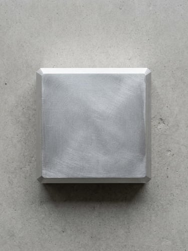 Top-down macro shot of a single raw aluminum paperweight on a smooth concrete surface, soft diffused north-facing daylight, quiet shadows