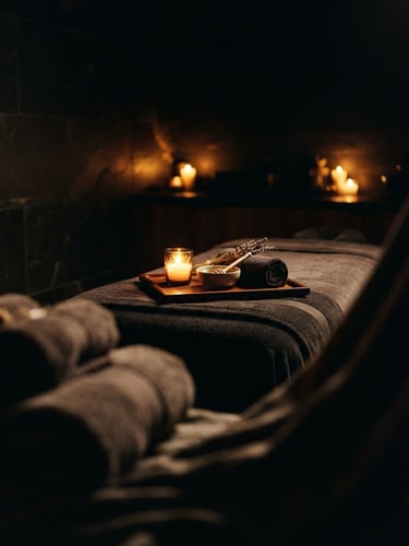 Low-key dramatic chiaroscuro shot of a candle-lit private massage suite, warm golden glow reflecting off dark polished stone, soft-focus linen in foreground.