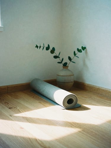 Soft-focus film photograph of a quiet studio corner, warm morning sun casting long shadows on a light wood floor, a rolled linen yoga mat resting beside a single green branch in a ceramic vase.